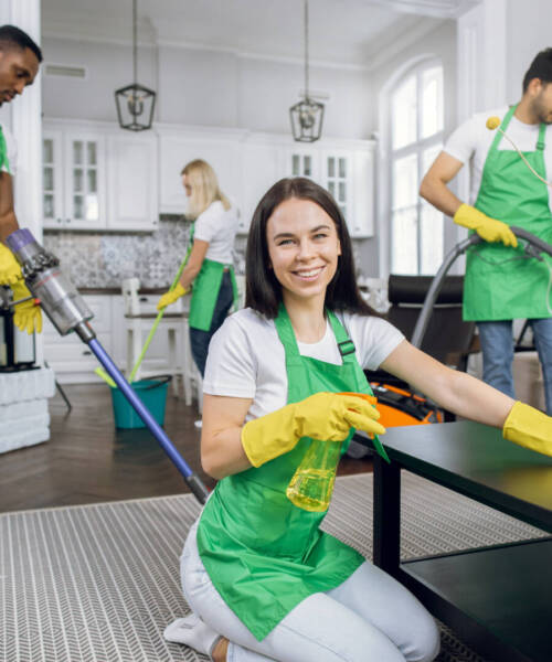 Pretty smiling Caucasian woman in green apron and yellow gloves, wiping table with microfiber cloth and detergents and smiling to camera. Cleaning team tidying up on the background